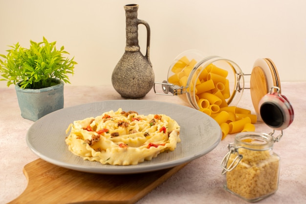 Итальянская паста, вид спереди, вкусная еда с приготовленными овощами и небольшими кусочками мяса внутри серой тарелки вместе с цветком и сырой пастой на розовом