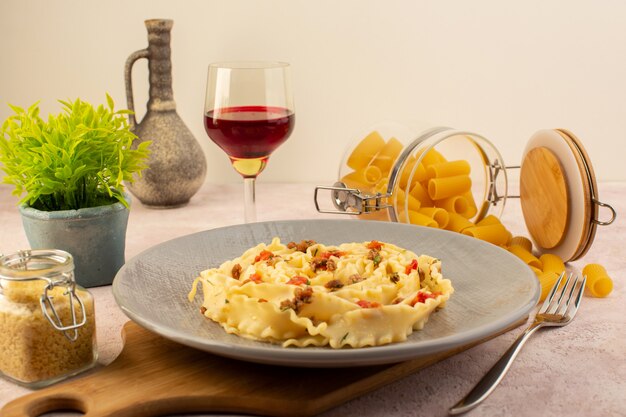 A front view italian pasta tasty meal with cooked vegetables and little meat slices inside grey plate along with flower and raw pasta on pink