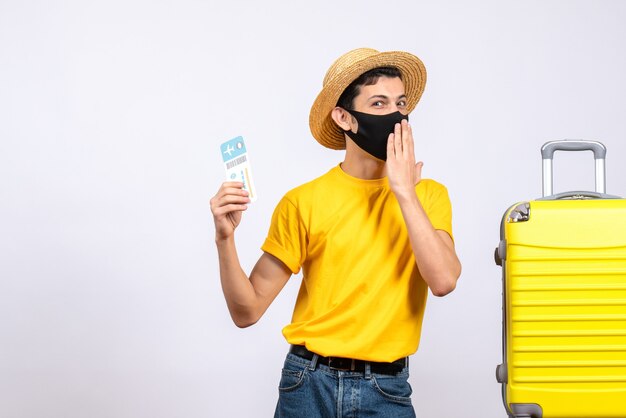 Front view joyful tourist in yellow t-shirt standing near yellow suitcase holding up travel ticket