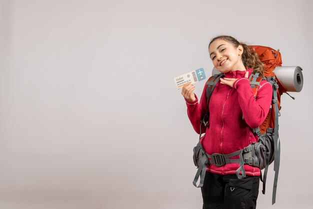 Front view of joyful young traveller with big backpack holding up travel ticket on grey wall