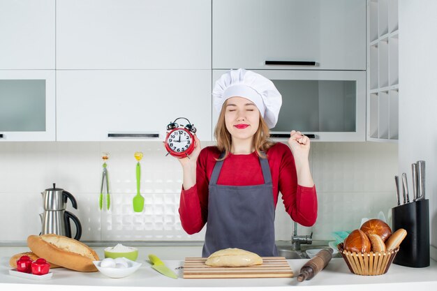 キッチンで赤い目覚まし時計を保持しているクック帽子とエプロンの正面図うれしそうな若い女性