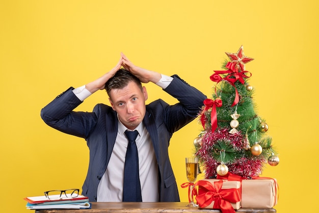 Vista frontale dell'uomo senza gioia che fa la casa sul tetto con le sue mani seduto al tavolo vicino all'albero di natale e regali su giallo