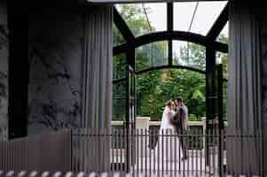 Foto gratuita vista frontale della finestra ampia e trasparente con porta aperta al di fuori della quale gli sposi novelli posano e si abbracciano sul balcone durante il giorno del loro matrimonio