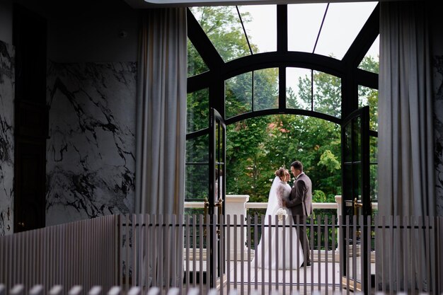 Вид спереди на большое и прозрачное окно с открытой дверью, за которым молодожены позируют и обнимаются на балконе в день свадьбы