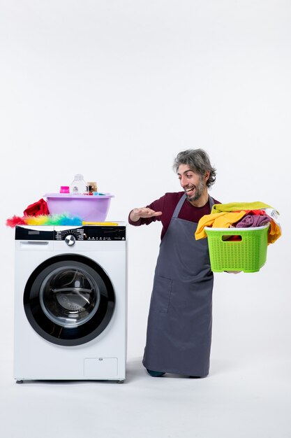 Front view laughing housekeeper man kneeing near washer holding laundry basket on white background