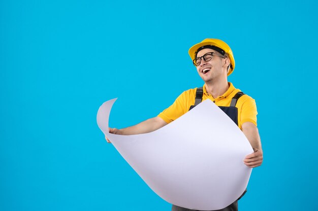 Front view laughing male builder in uniform with paper plan on blue 