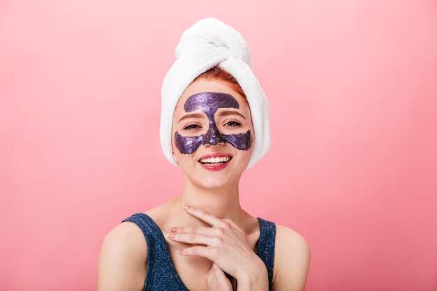 Front view of laughing woman in towel and mask isolated on pink background. Happy girl doing face treament with smile.