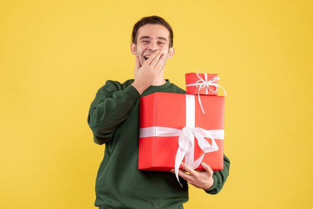 黄色の上に立っているクリスマスプレゼントと若い男を笑っている正面図