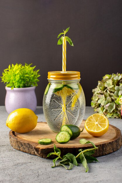Вид спереди лимонный коктейль свежий прохладный напиток внутри стеклянной чашки нарезанные лимоны огурцы солома на деревянный стол и серый фон коктейль напиток фрукты