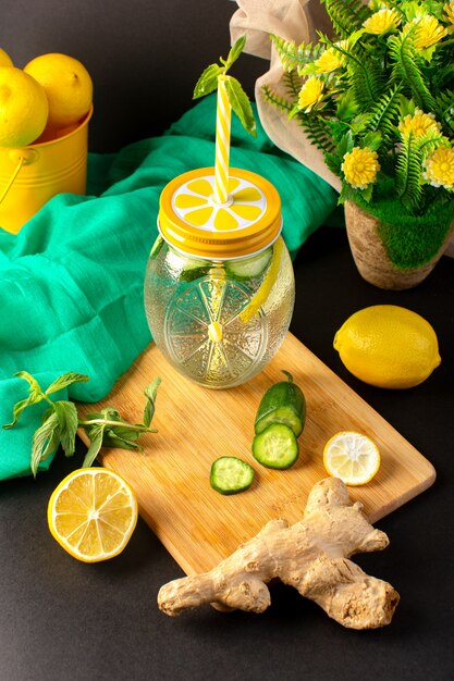 A front view lemon cocktail fresh cool drink inside glass cup sliced and whole lemons cucumbers along with flowers on the dark background cocktail drink fruit