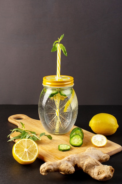 Free photo a front view lemon cocktail fresh cool drink inside glass cup sliced and whole lemons cucumbers along with flowers straw on the dark background cocktail drink fruit