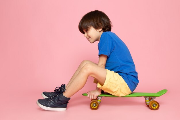 A front view little boy in blue t-shirt and yellow jeans riding green skateboard on the pink space