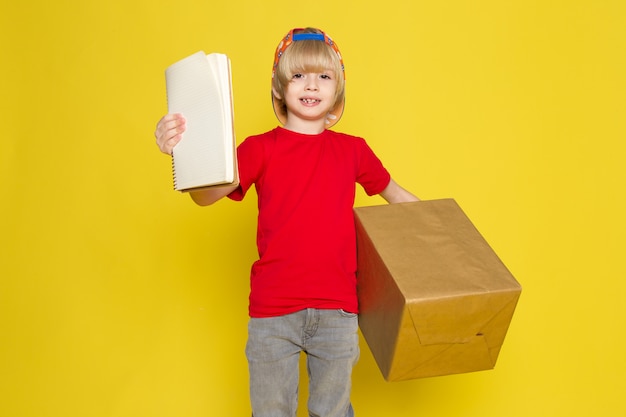 A front view little boy in red t-shirt colorful cap and grey jeans holding box on the yellow background