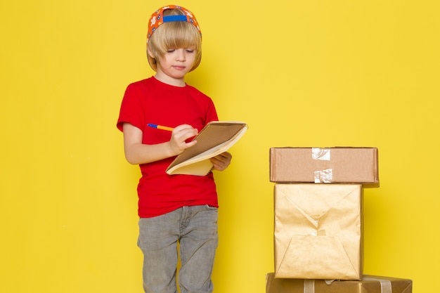 A front view little boy in red t-shirt colorful cap and grey jeans holding box on the yellow background