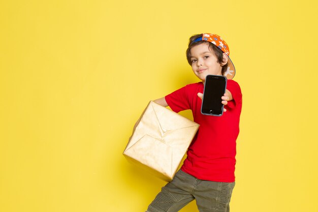 A front view little boy in red t-shirt colorful cap and khaki trousers holding box on the yellow background