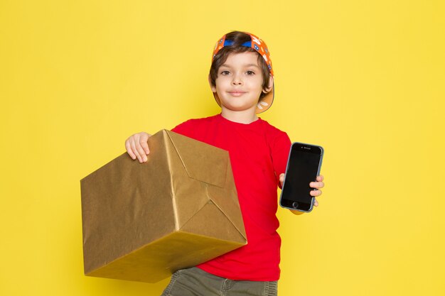 A front view little boy in red t-shirt colorful cap and khaki trousers holding box on the yellow background
