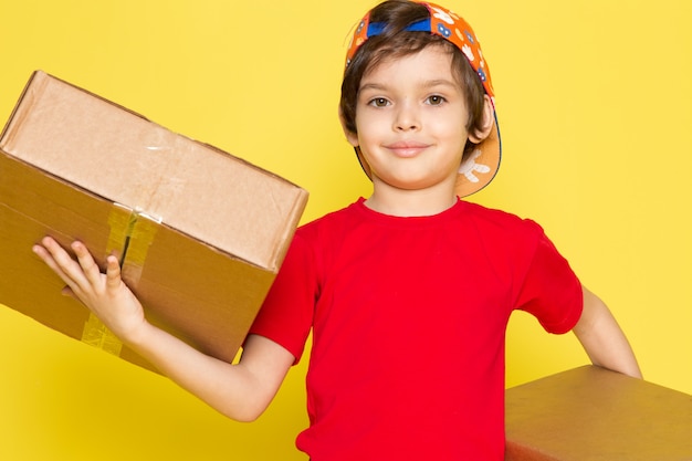 A front view little boy in red t-shirt colorful cap and khaki trousers holding box on the yellow background