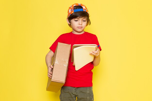 A front view little boy in red t-shirt colorful cap and khaki trousers holding box on the yellow background