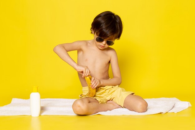 вид спереди маленький мальчик в солнцезащитные очки, очищающий банан на желтом