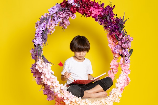 Free photo a front view little boy in white t-shirt reading book on the flower made stand on the yellow space