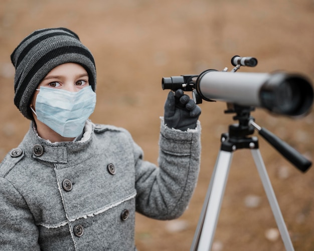 Front view little boy with medical mask using a telescope