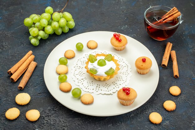 Front view little cake with cream and green grapes inside plate along with cookies cinnamon and tea on the dark surface fruit sweet 