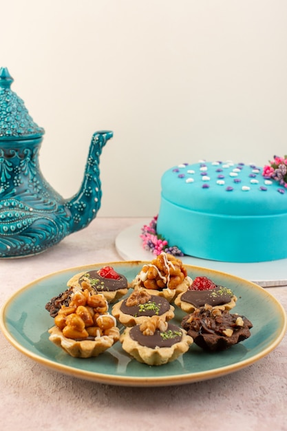 A front view little chocolate cakes inside plate with blue birthday cake on the pink desk