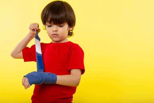 Вид спереди маленький милый мальчик, связывая его руку с синей тканью в красной футболке на желтой стене