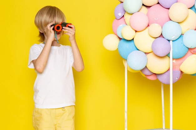 front view little cute boy in white t-shirt along with colorful air balloons on yellow desk