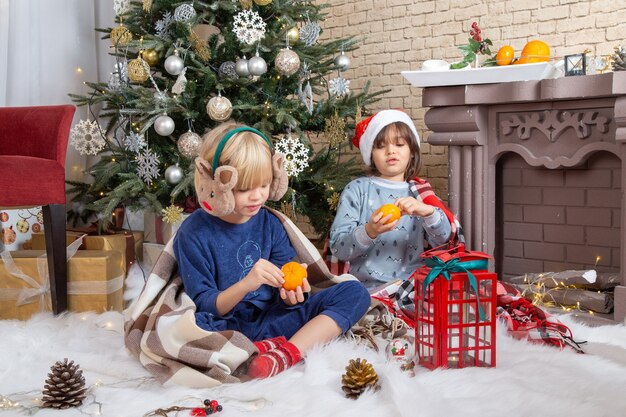 Front view little cute boys sitting around xmas tree and presents in their house eating tangerine kid color new year photo childhood christmas