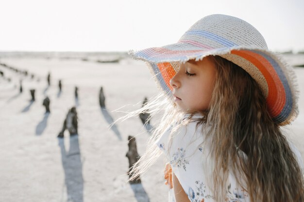 Front view of a little girl  with blonde hair dressed in the hat on the beach with closed eyes