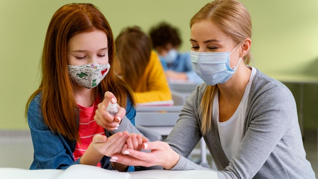Front view of little girl with medical mask getting hand sanitizer from teacher