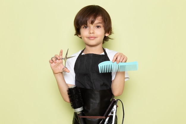 Un bambino adorabile del piccolo parrucchiere di vista frontale nella spazzola e nelle forbici della tenuta del mantello nero