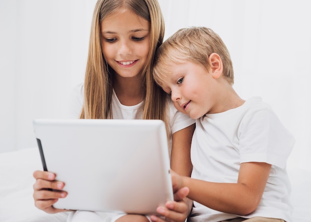 Front view little siblings staying in bed while looking at their tablet