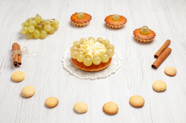 Вид спереди маленькие вкусные пирожные с виноградом и печеньем на белом столе, торт, бисквит, сладкое десертное печенье