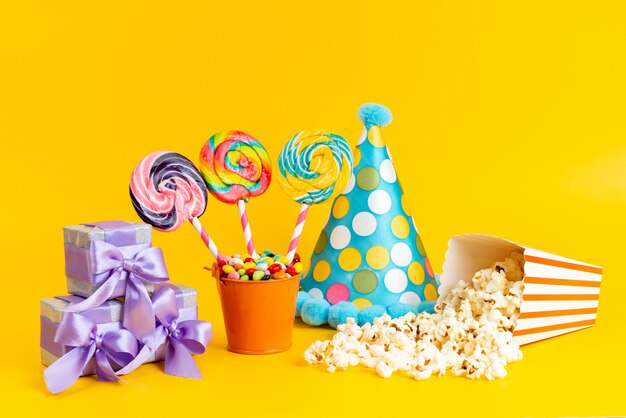 A front view lollipops and popcorn along with blue cap purple gift boxes and candies on yellow