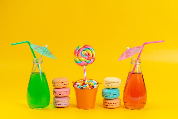 A front view macarons and cocktails colored with candies and lollipop on yellow, color cake biscuit rainbow