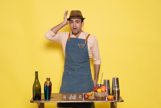 Вид спереди мужчина-бармен перед столом с напитками и шейкерами на светло-желтой стене пить ночной алкогольный бар цветной клуб