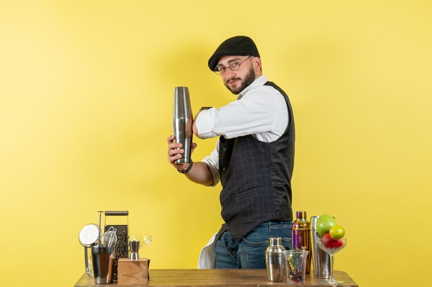 Вид спереди мужчина-бармен перед столом с шейкерами, делающий напиток на желтой стене, алкогольный ночной клуб молодежных напитков
