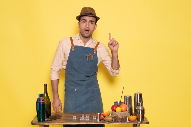 Front view male bartender standing in front of desk with drinks and fruits posing on the yellow wall drinks night club bar male