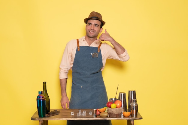 Front view male bartender standing in front of desk with drinks and fruits smiling on yellow wall night club bar male drink