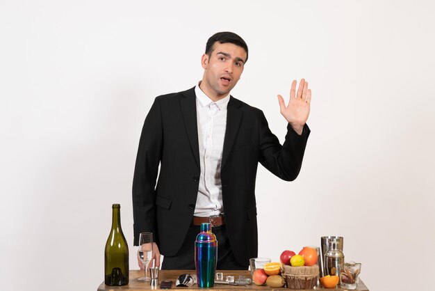 白い壁に飲み物とシェーカーを持ってテーブルの前に立っている正面図男性バーテンダー飲み物アルコール男性ナイトバークラブ