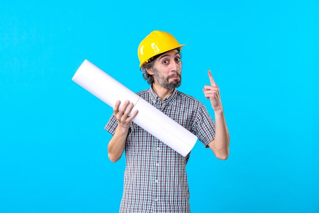 Front view male builder in helmet holding plan on the blue background engineer job constructor skyscraper building color architecture