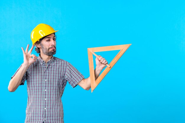 Front view male builder in helmet holding triangle wooden figure on blue background architecture job constructor design color building