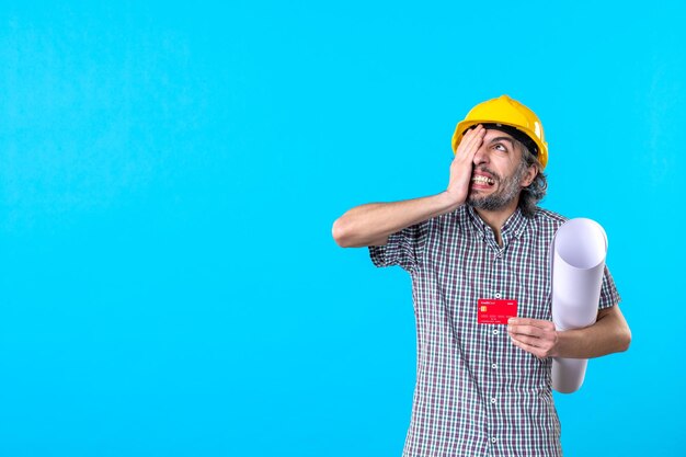 Вид спереди мужской строитель, держащий план и банковскую карту на синем фоне, деньги, дизайн, конструктор, строительство, работа, работа, цвета, архитектура