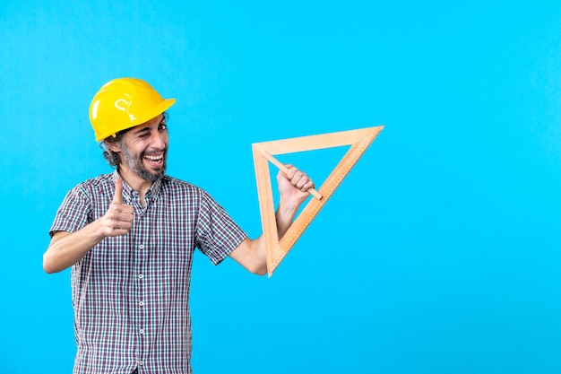 Бесплатное фото Вид спереди мужчина-строитель в шлеме, держащий треугольную деревянную фигуру на синем фоне, инженер архитектуры, конструктор, дизайн, цвета, строительные работы