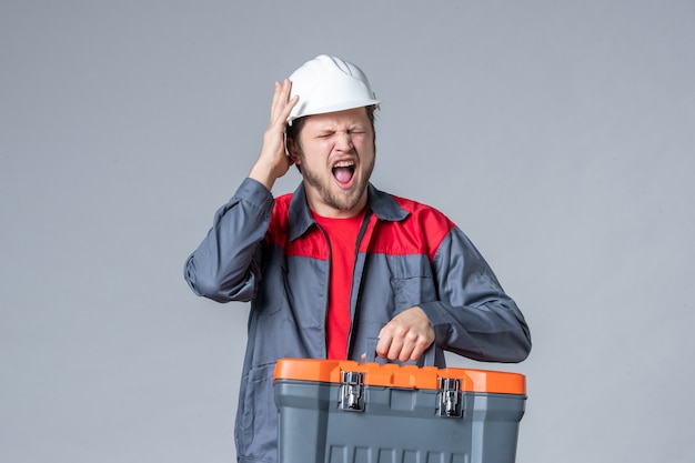 Бесплатное фото Мужчина-строитель в униформе, держащий чемодан с инструментами на сером фоне, вид спереди