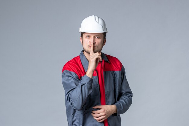 мужчина-строитель в форме и шлеме, просящий молчать на сером фоне, вид спереди