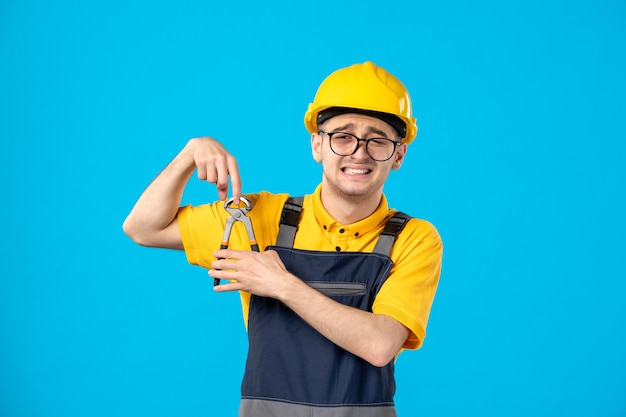 Мужчина-строитель в униформе и шлеме, режущий палец плоскогубцами на синем, вид спереди