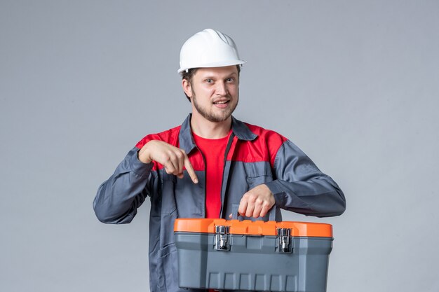 мужчина-строитель в форме и шлеме, держащий чемодан с инструментами на сером фоне, вид спереди